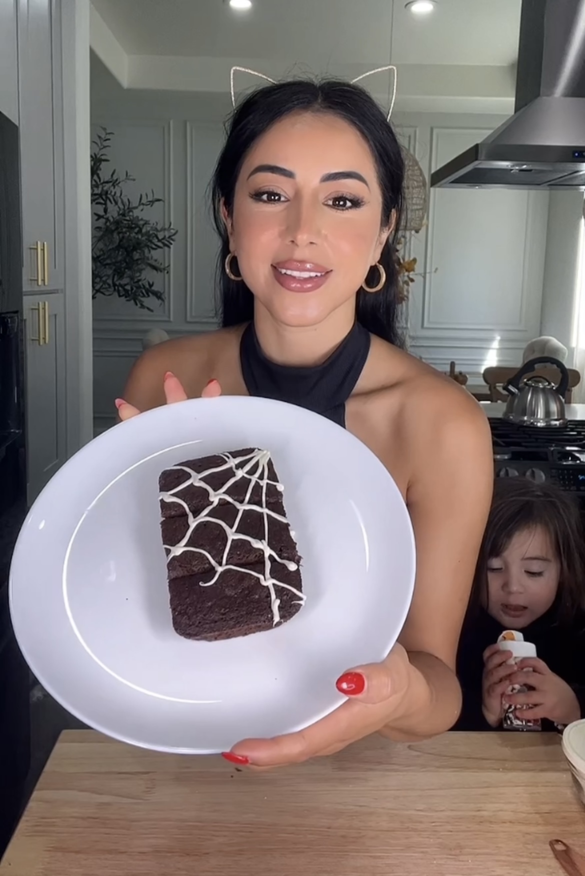 woman wearing halloween cat ears holding a plate with chocolate brownies decorated with a white chocolate spider web
